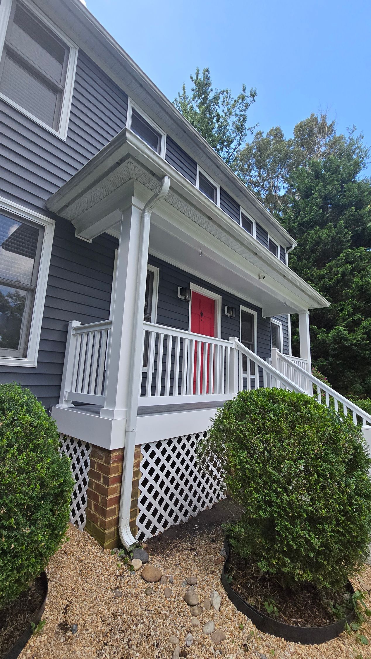 Gray house front porch with white railings, red door, and trimmed bushes beside stone landscaping