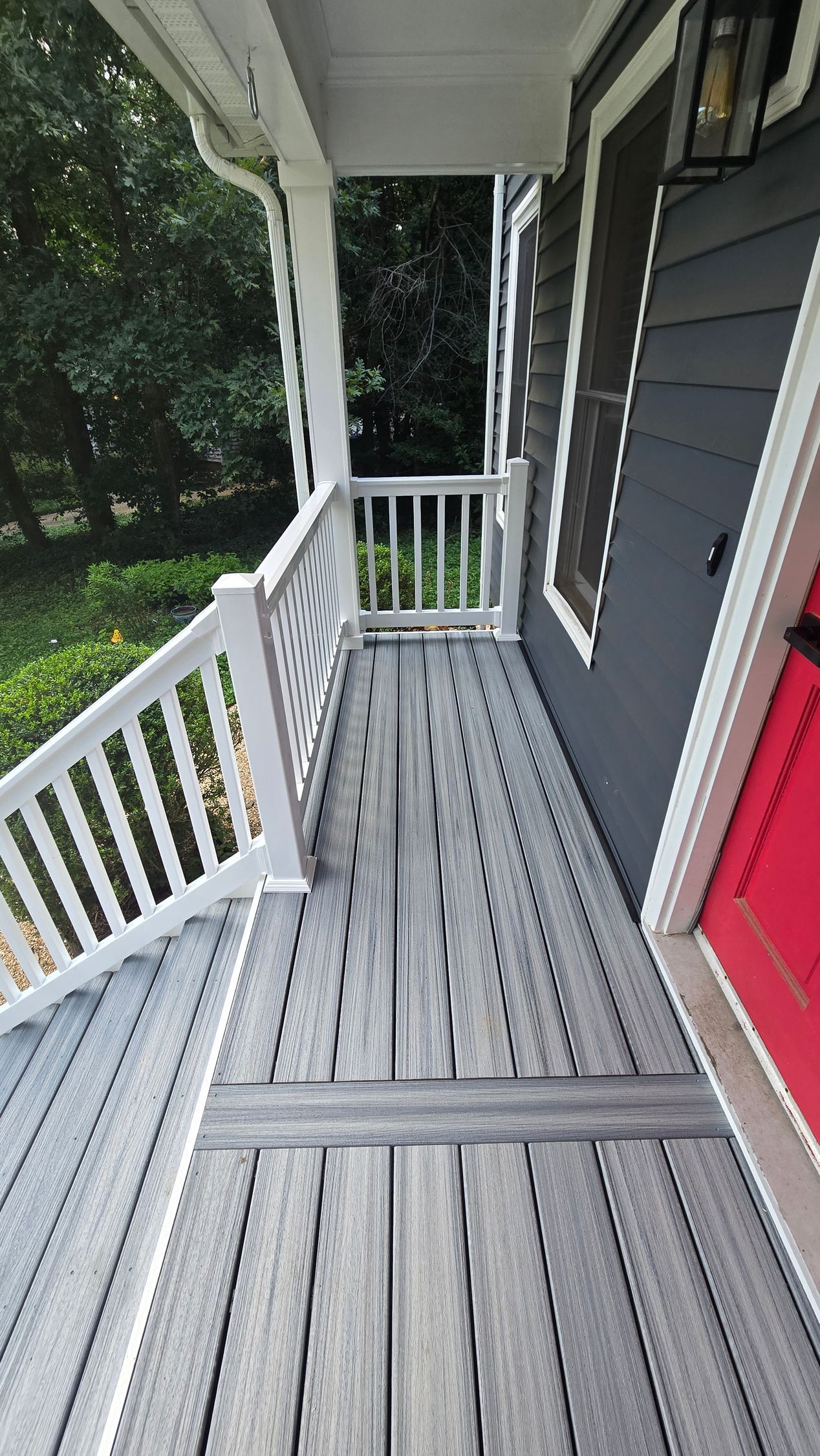 Front porch with gray wooden floor, white railings, dark siding, and a red front door.