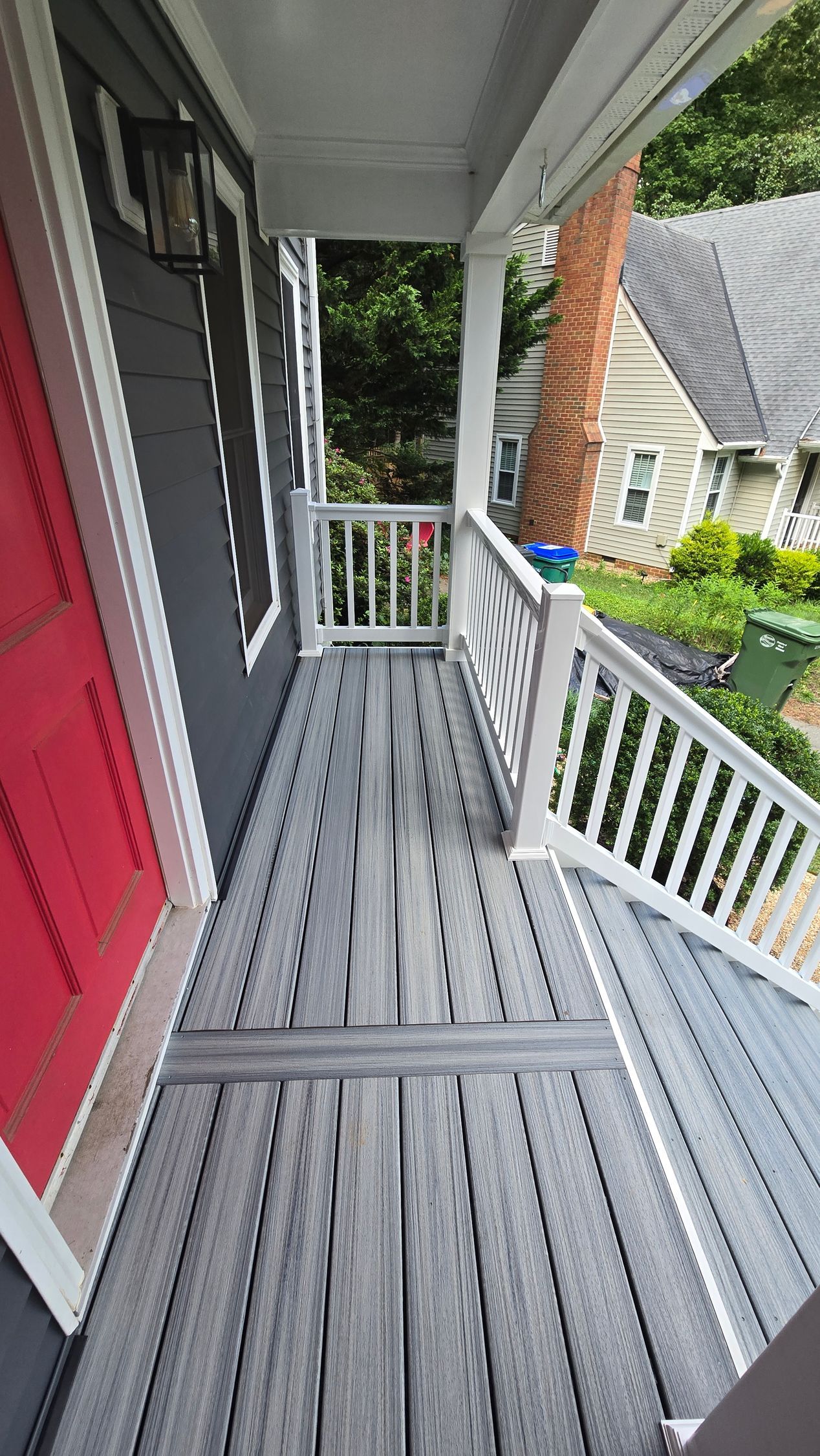 Narrow gray front porch with white railing and red wall, overlooking a yard and neighboring houses.