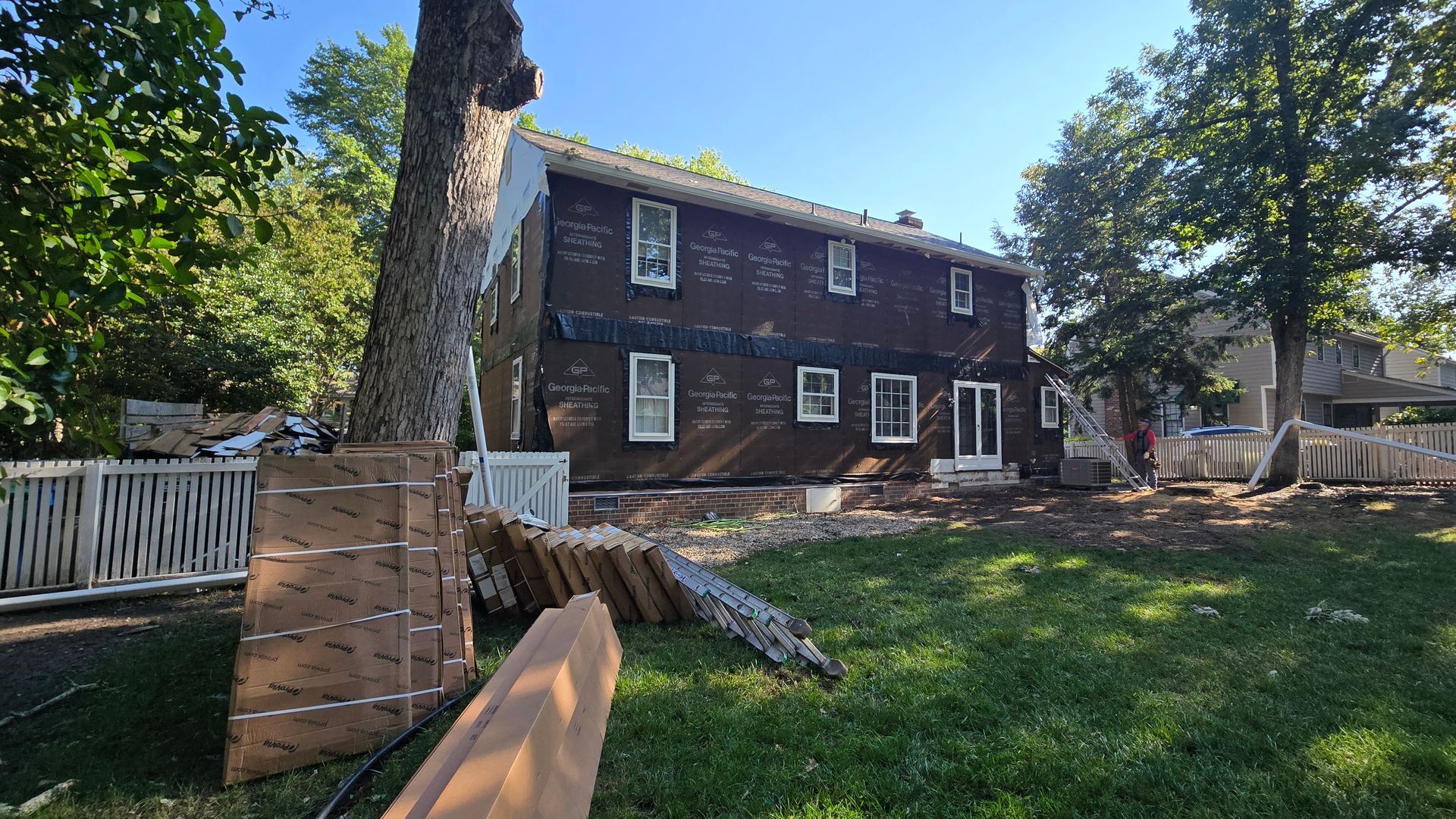 Two-story dark brown house with white trim, large tree in foreground, and sunny green yard