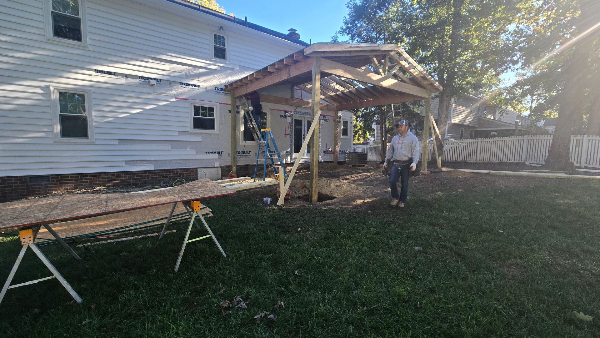 Backyard patio under construction beside a white house, with sawhorses and a person near the framed roof.