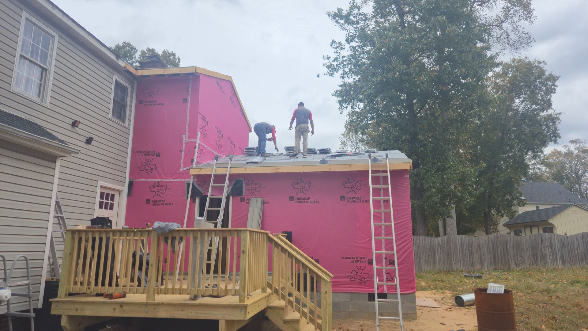 Two workers on the roof of a pink house, with ladders and a wooden deck in the backyard.