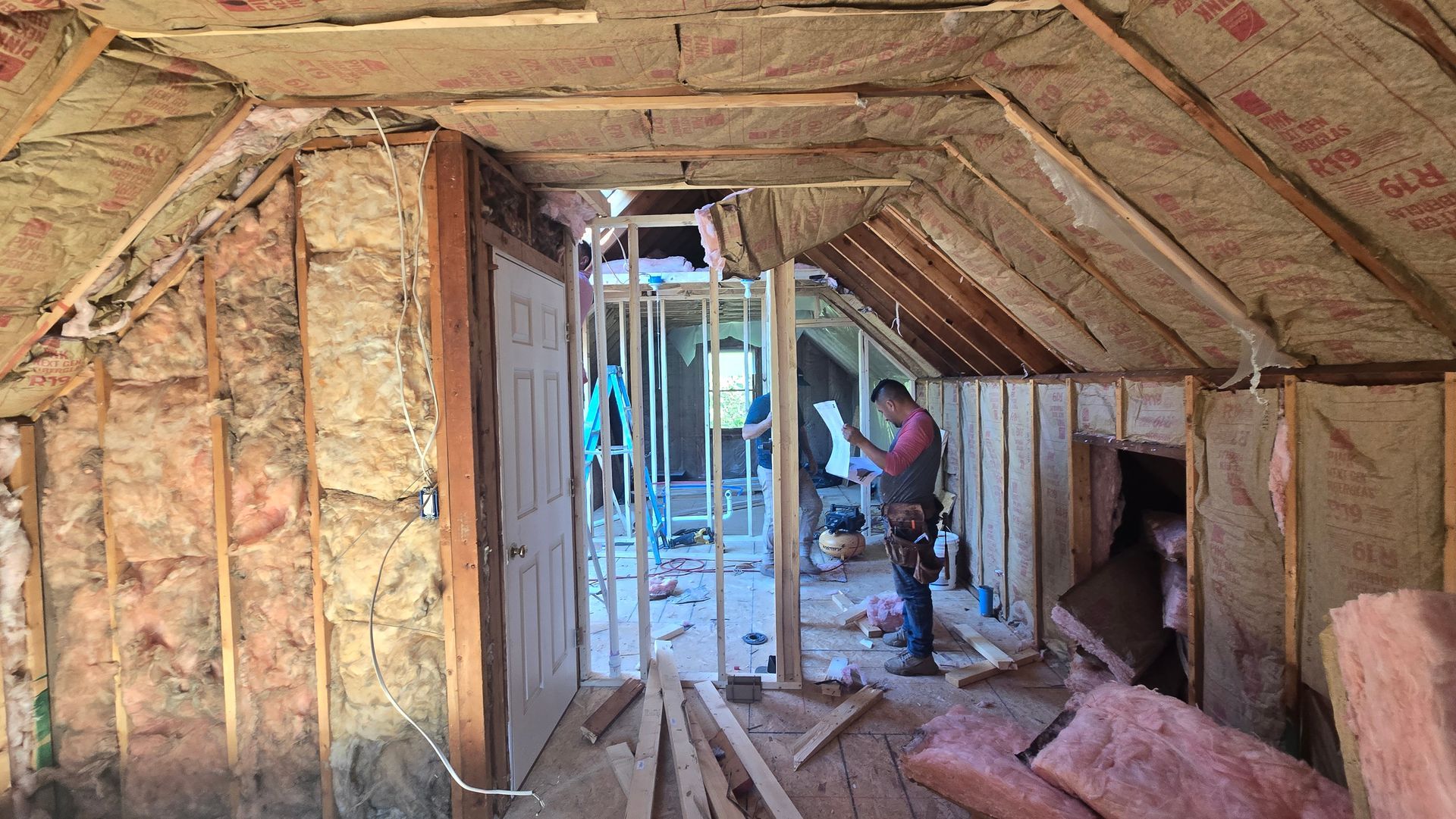 Attic under construction with exposed insulation and a worker using tools near the center window opening