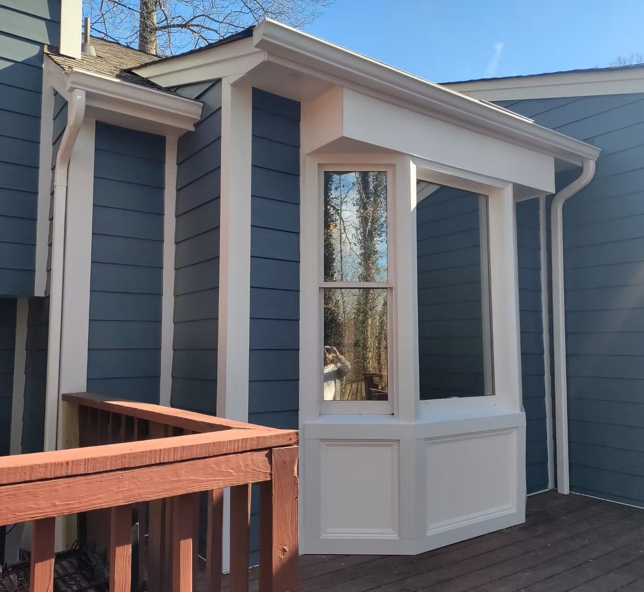 Blue house entry with a small white bay window and wood deck railing in daylight
