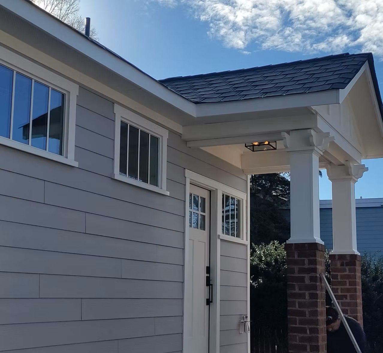 Front porch of a gray house with white trim, brick column, and double doors under a blue sky.