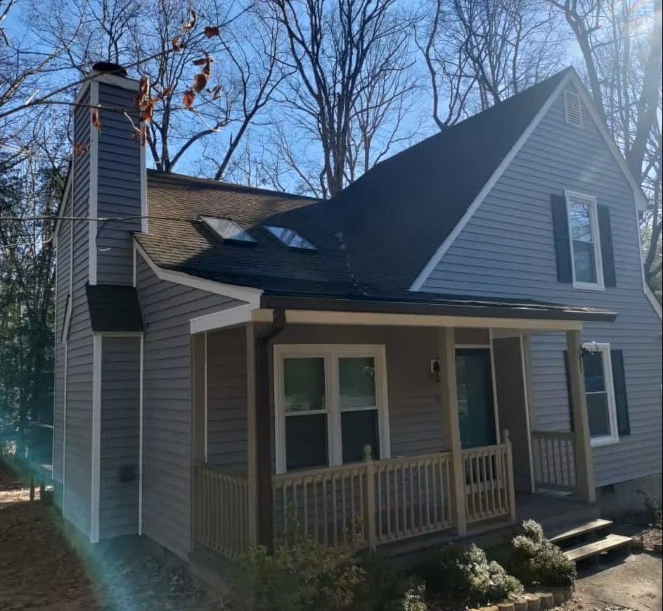 Gray house with white trim, steep roof, front porch, and chimney surrounded by leafless trees.