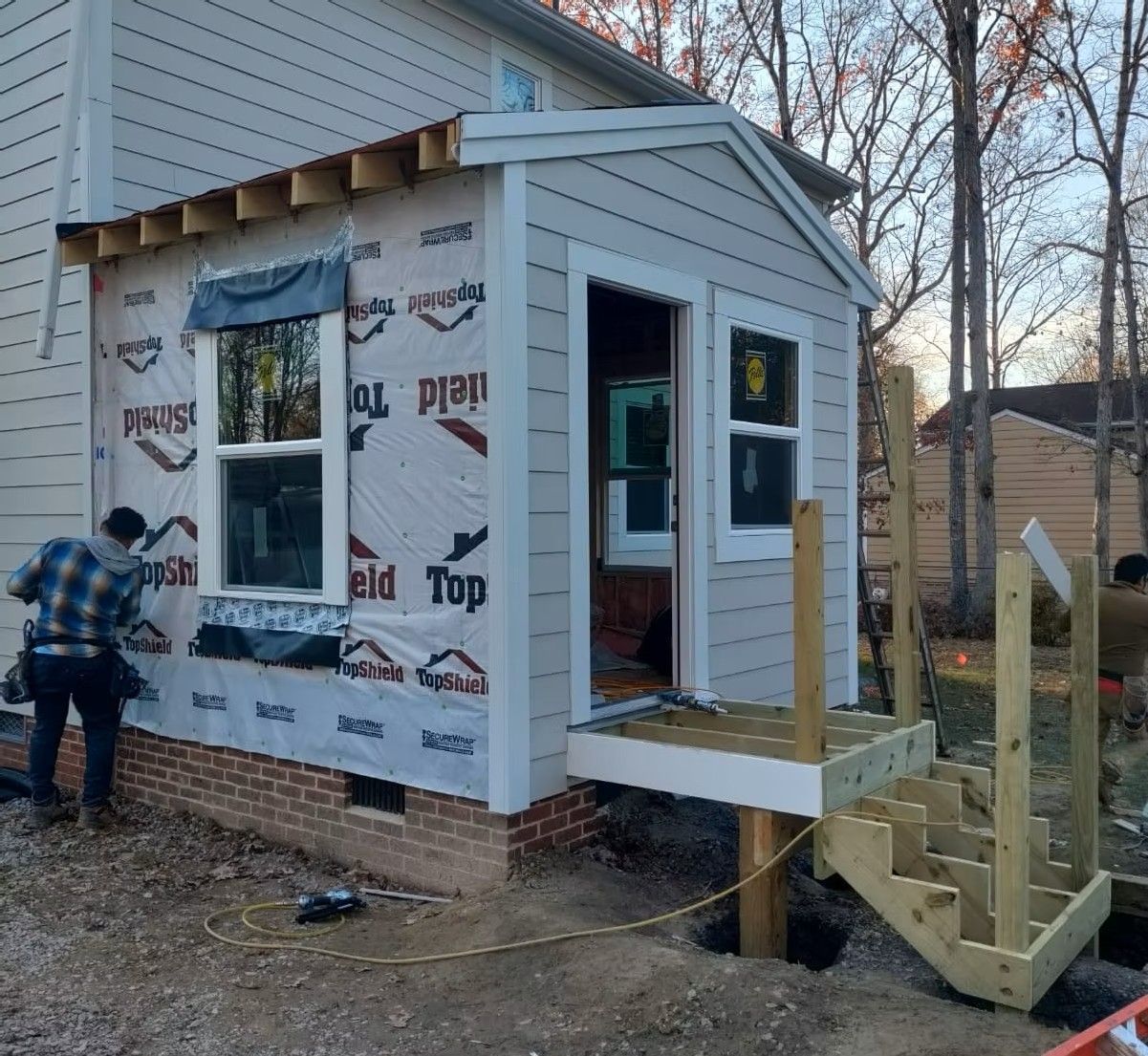 House under construction with porch framing, house wrap, and a worker near the side wall
