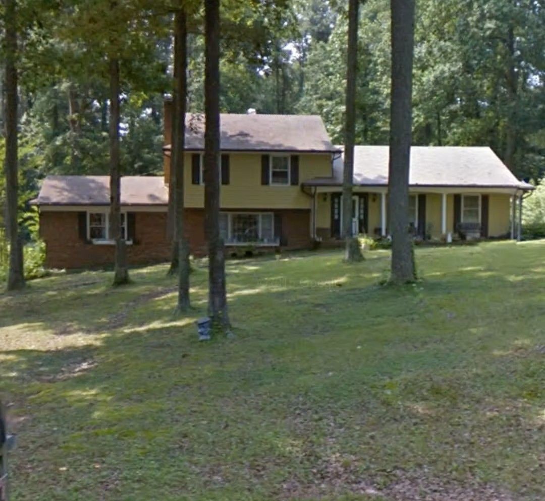 Two-story tan house with a porch and carport among tall trees on a grassy lawn