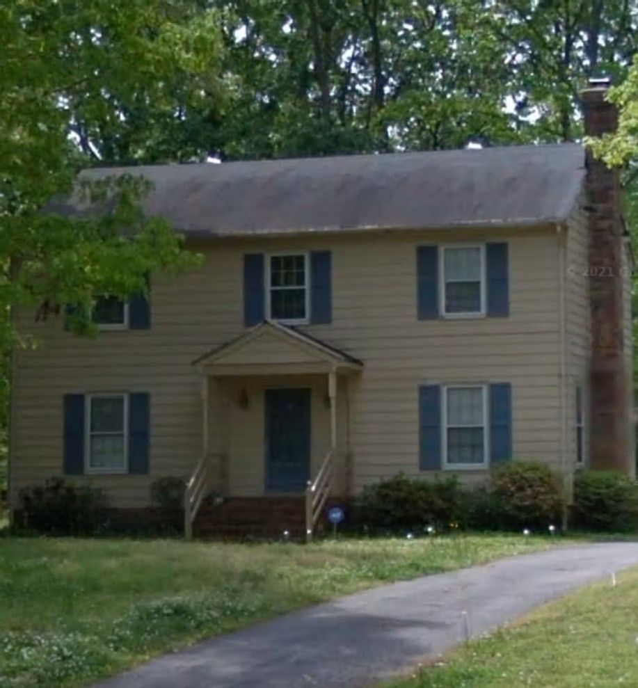 Two-story beige house with blue shutters, front porch, brick chimney, and a driveway in a wooded yard