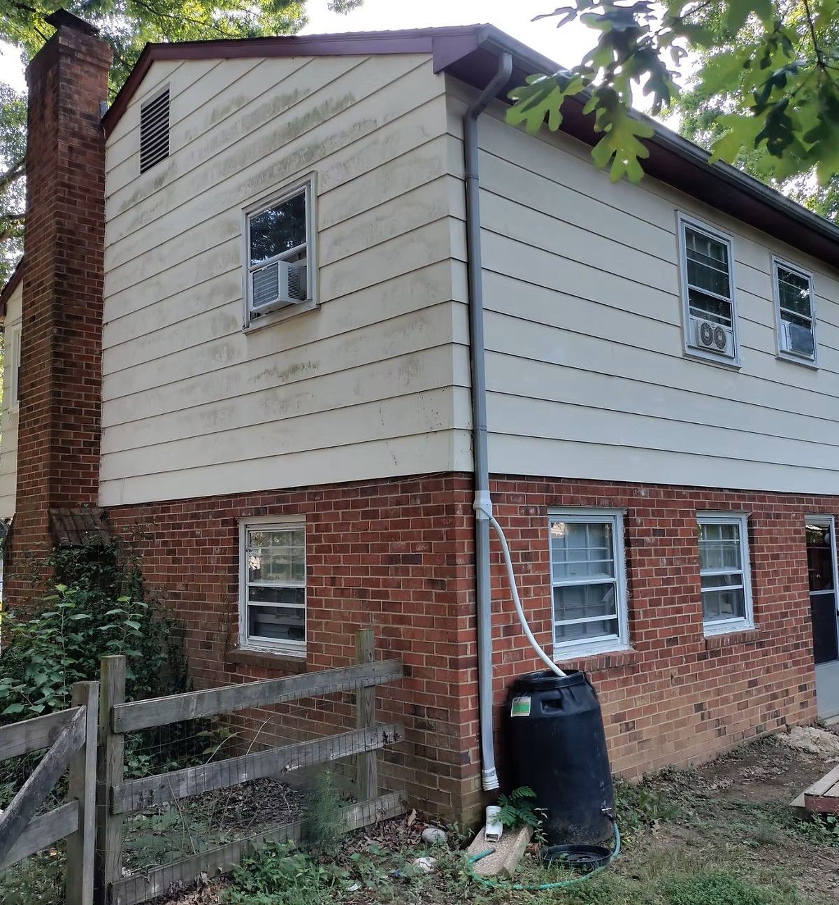 Two-story house with red brick lower level, beige siding above, and a black rain barrel by the downspout.