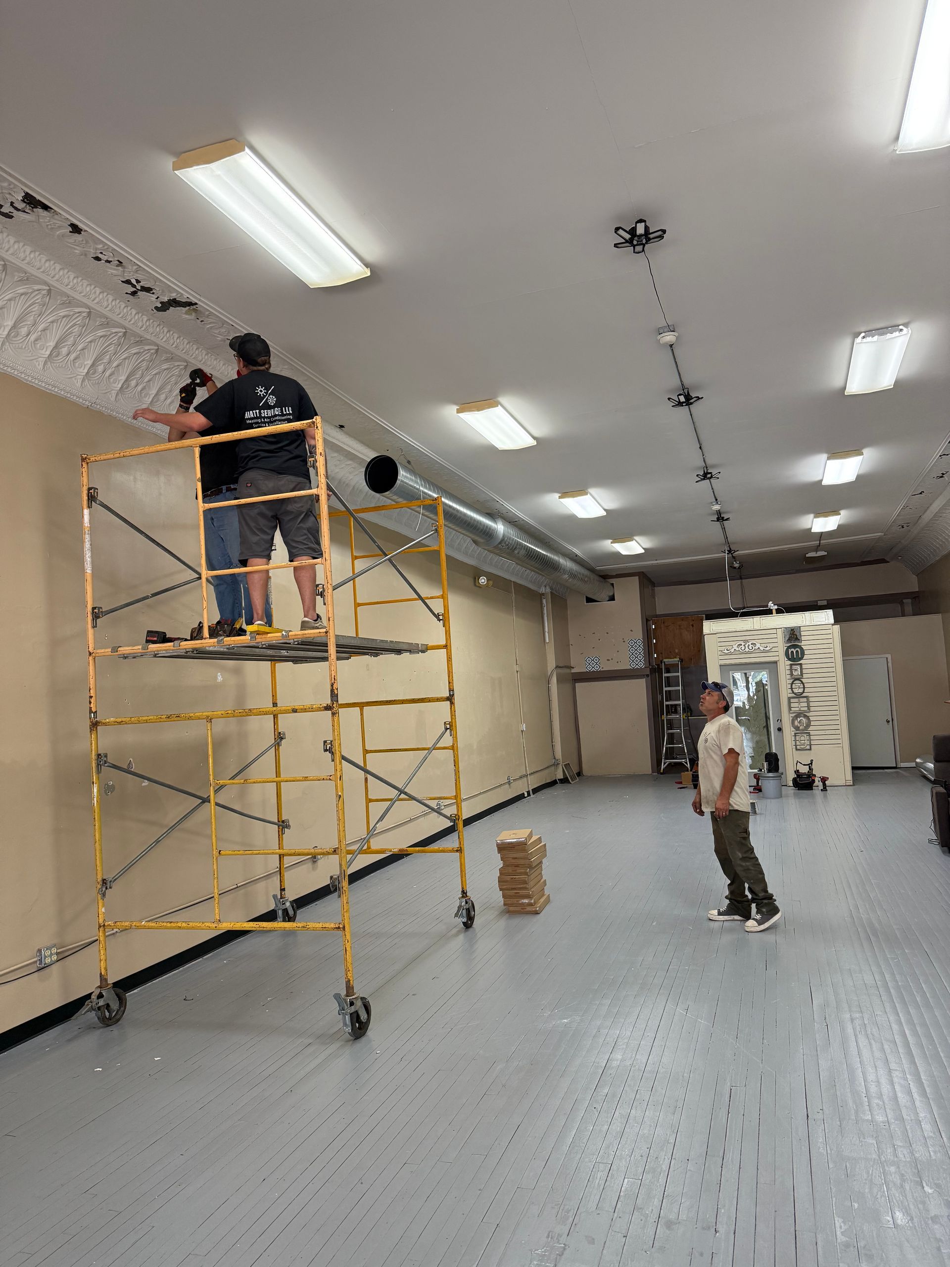 Two men working, one on a yellow scaffold. White ceiling with fluorescent lights.