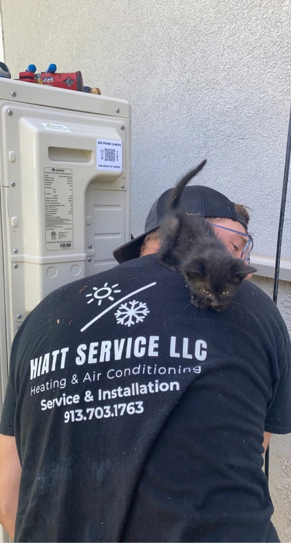 Man wearing a Matt Service LLC shirt working on HVAC unit, with a cat perched on his back.