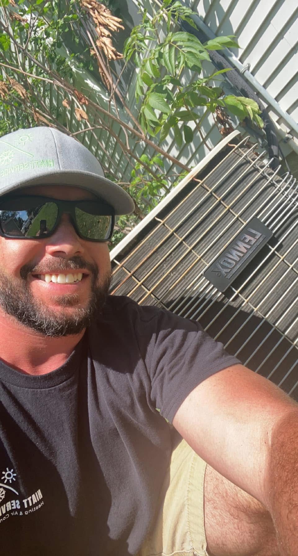 Man with sunglasses smiling next to an AC unit; outdoor setting.
