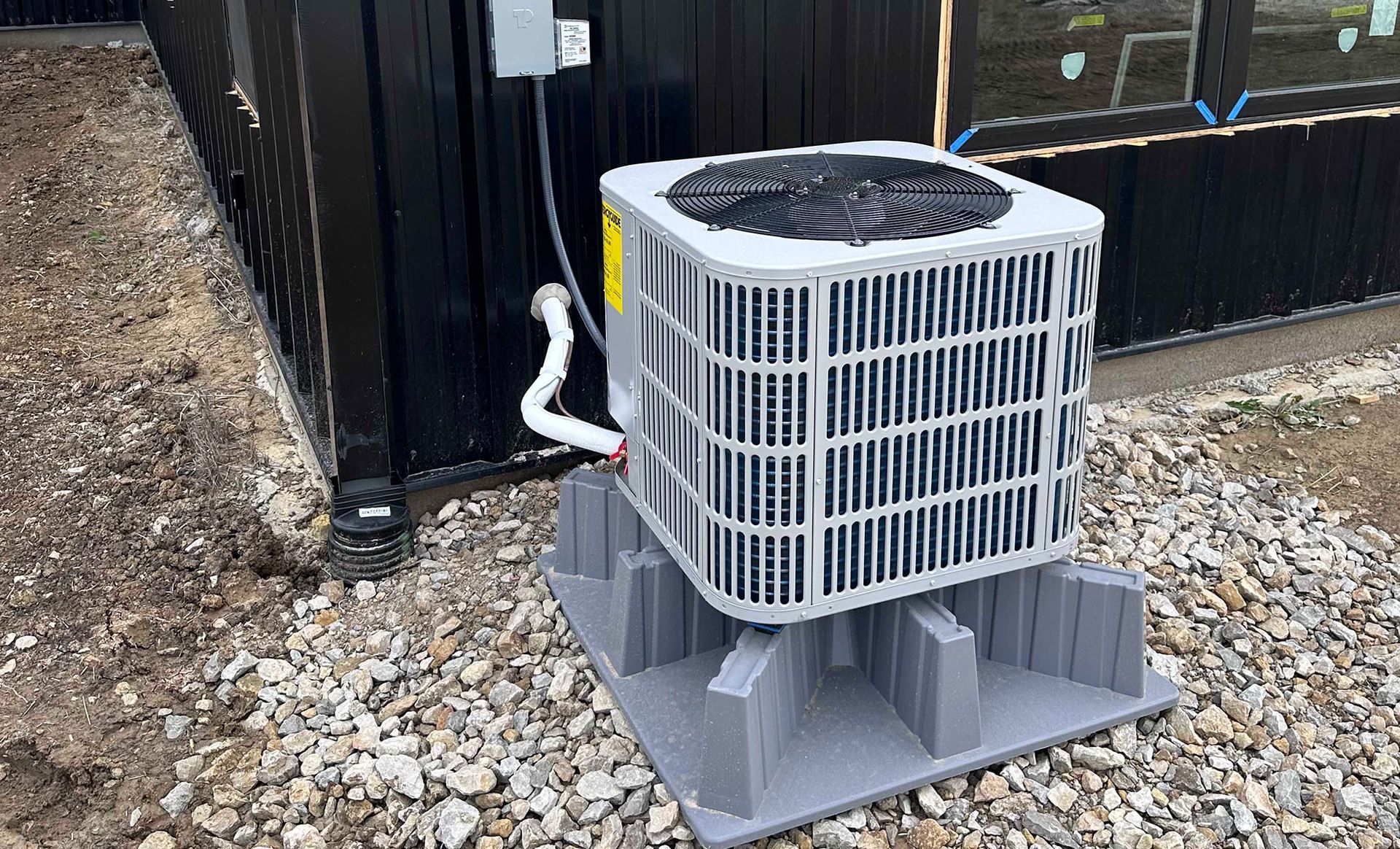 Air conditioning unit on gray base next to a black building, on gravel ground.