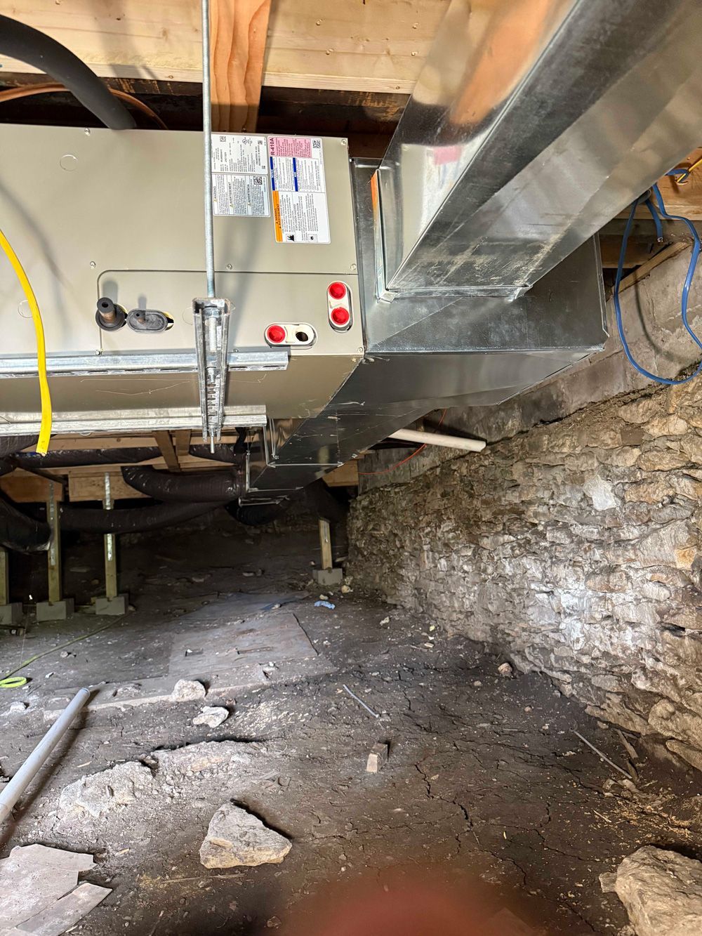 HVAC unit installed under a home, supported by wooden blocks over a dirt floor next to a stone wall.