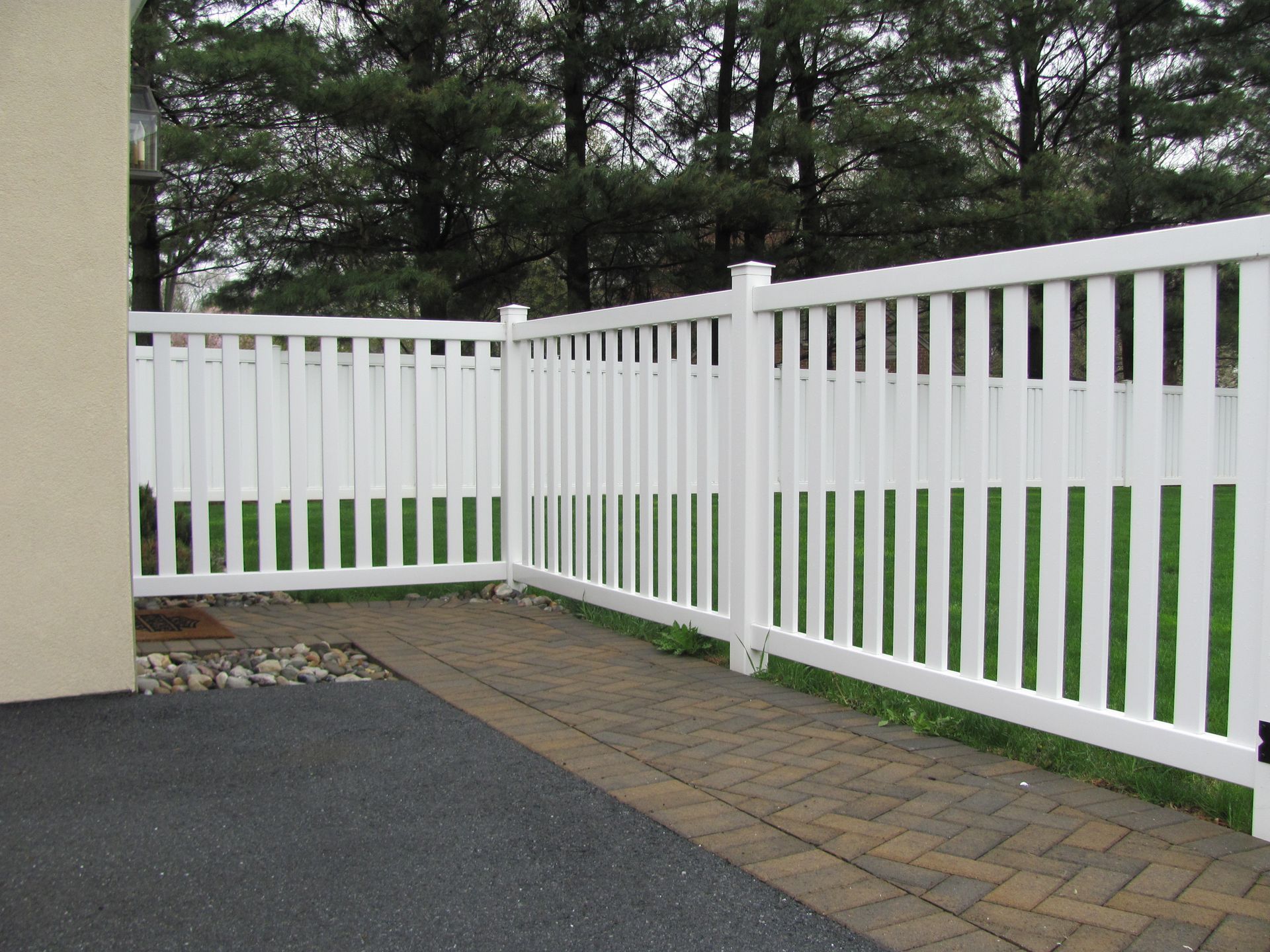 A white fence surrounds a brick walkway with trees in the background