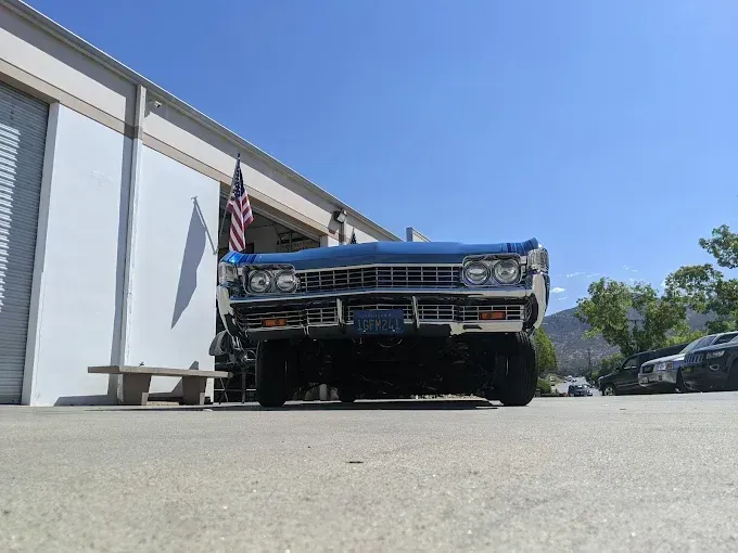 Blue classic car parked in front of a white building with an American flag on a sunny day.