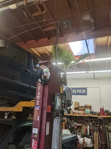 Car lifted in a repair shop, palm tree visible through skylight, work area cluttered.