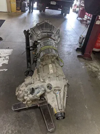 Large, dirty transmission on a metal stand in a repair shop, with a vehicle visible in the background.
