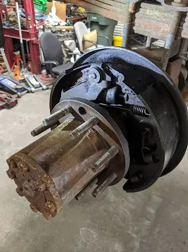 A rusty truck axle assembly with a black brake drum, in a cluttered workshop.