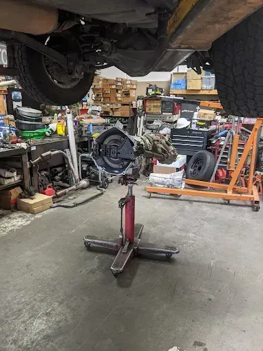 A car's undercarriage in a garage, a transmission on a stand in front of it.