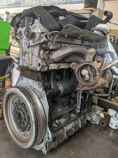 Car engine, gray and black, showing internal components.  Visible flywheel, exhaust manifold, and other parts. In a shop setting.