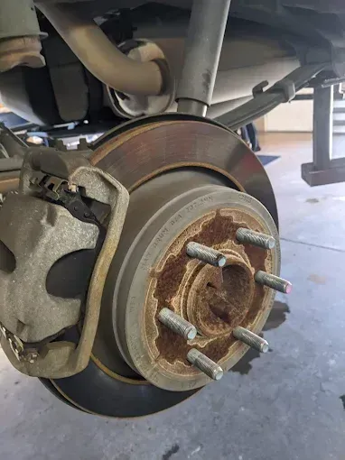 Car's rear brake disc and caliper assembly, showing rust on the rotor and hub, and exhaust system in the background.