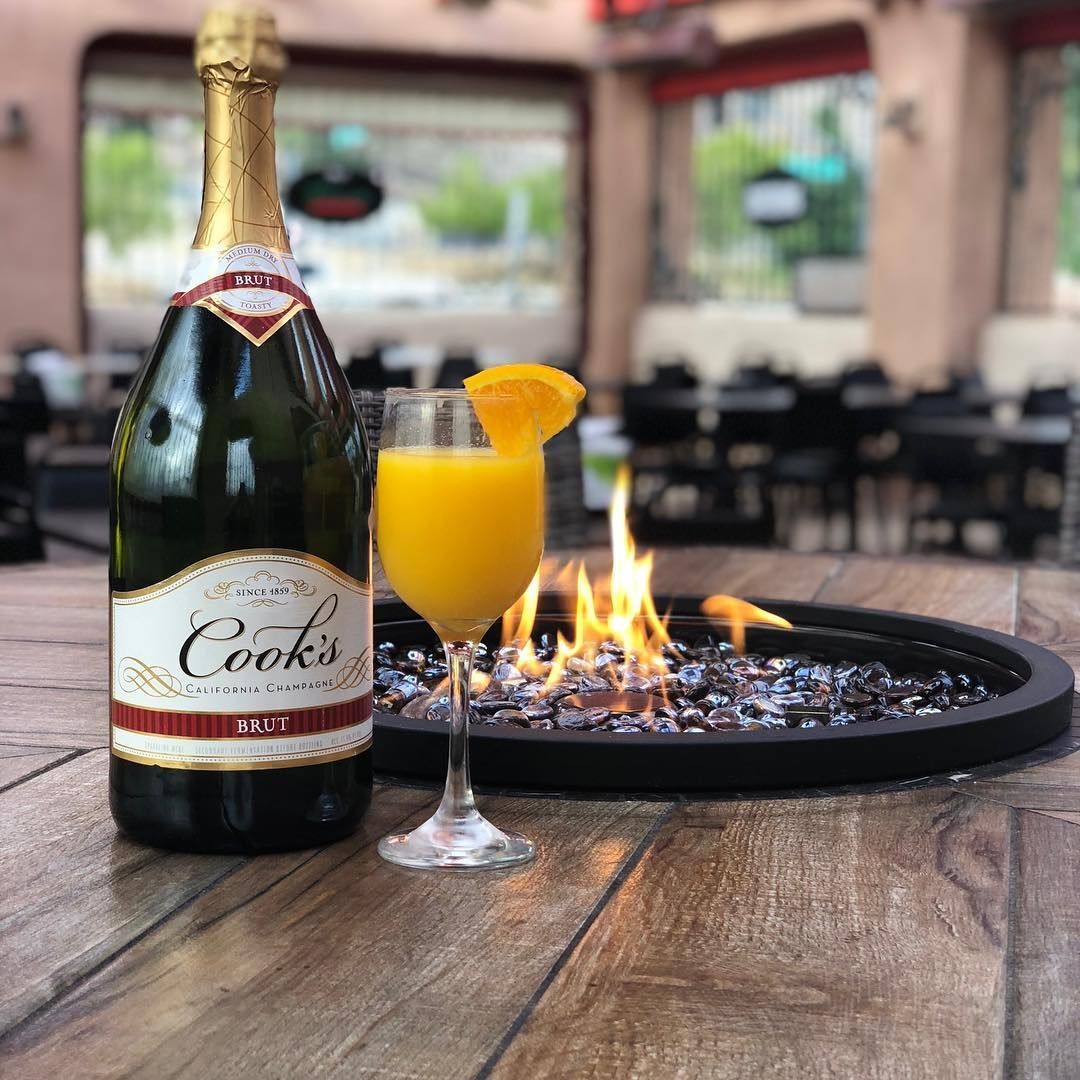 Bottle of Cook's champagne and mimosa with orange slice, next to a lit fire pit on a patio.