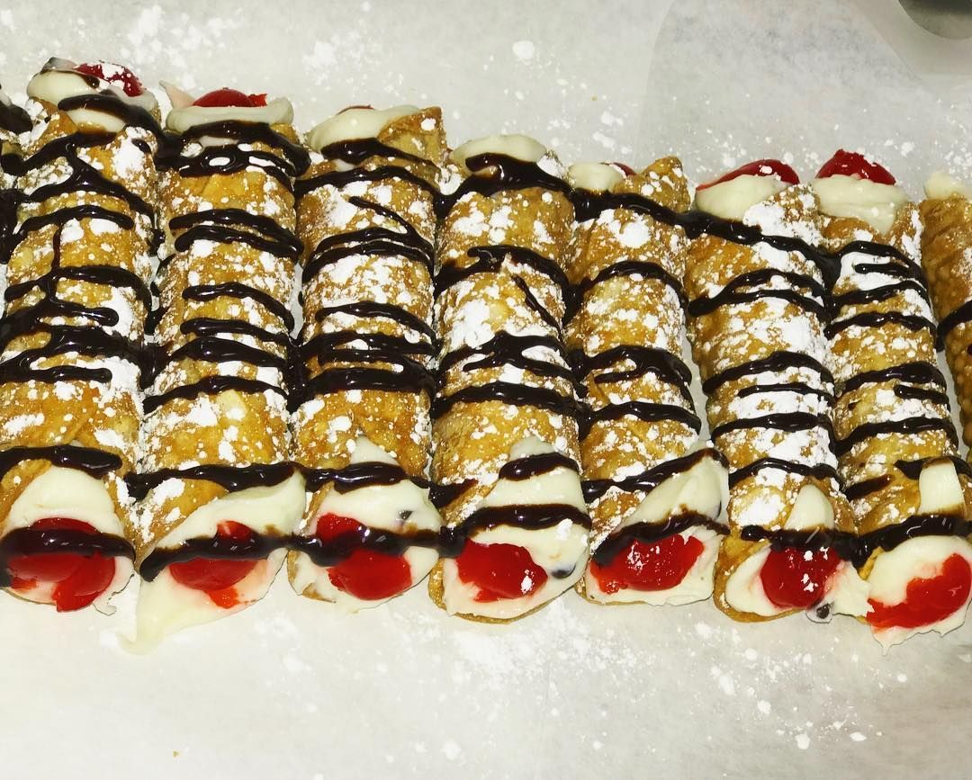 Cannoli pastries lined up, filled with white cream and red filling, drizzled with chocolate, dusted with powder.