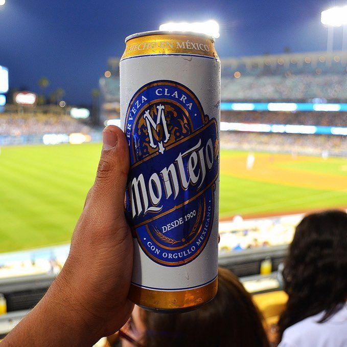 Person holding a can of Montejo beer at a baseball game, with a green field and stadium seating in the background.