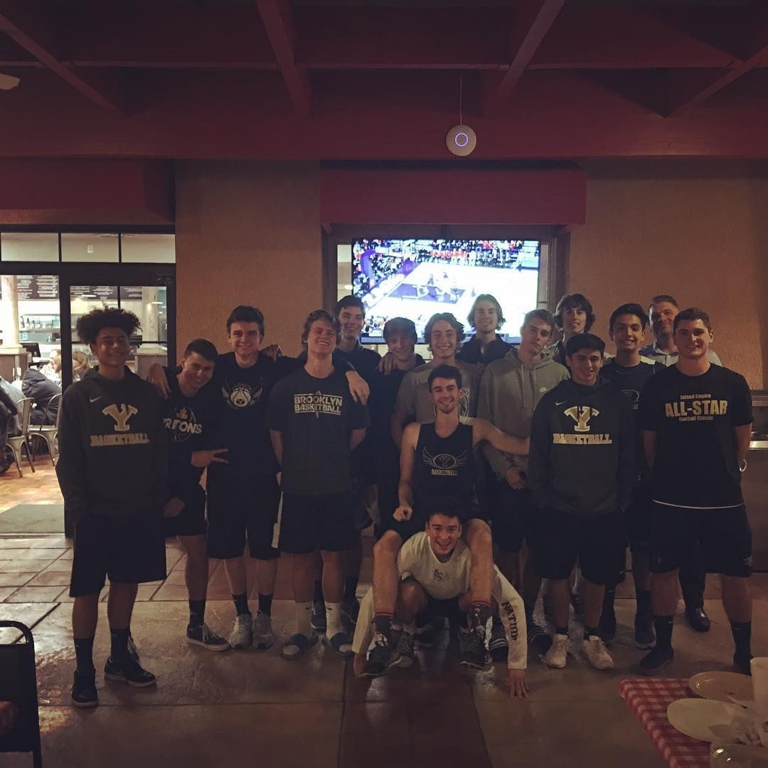 A group of teenage basketball players pose for a photo in a restaurant; some are wearing team gear.