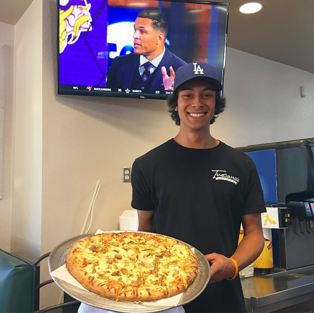 Man holding pizza, smiling, wearing a black t-shirt and a Los Angeles Dodgers hat. A TV plays a sports game in the background.