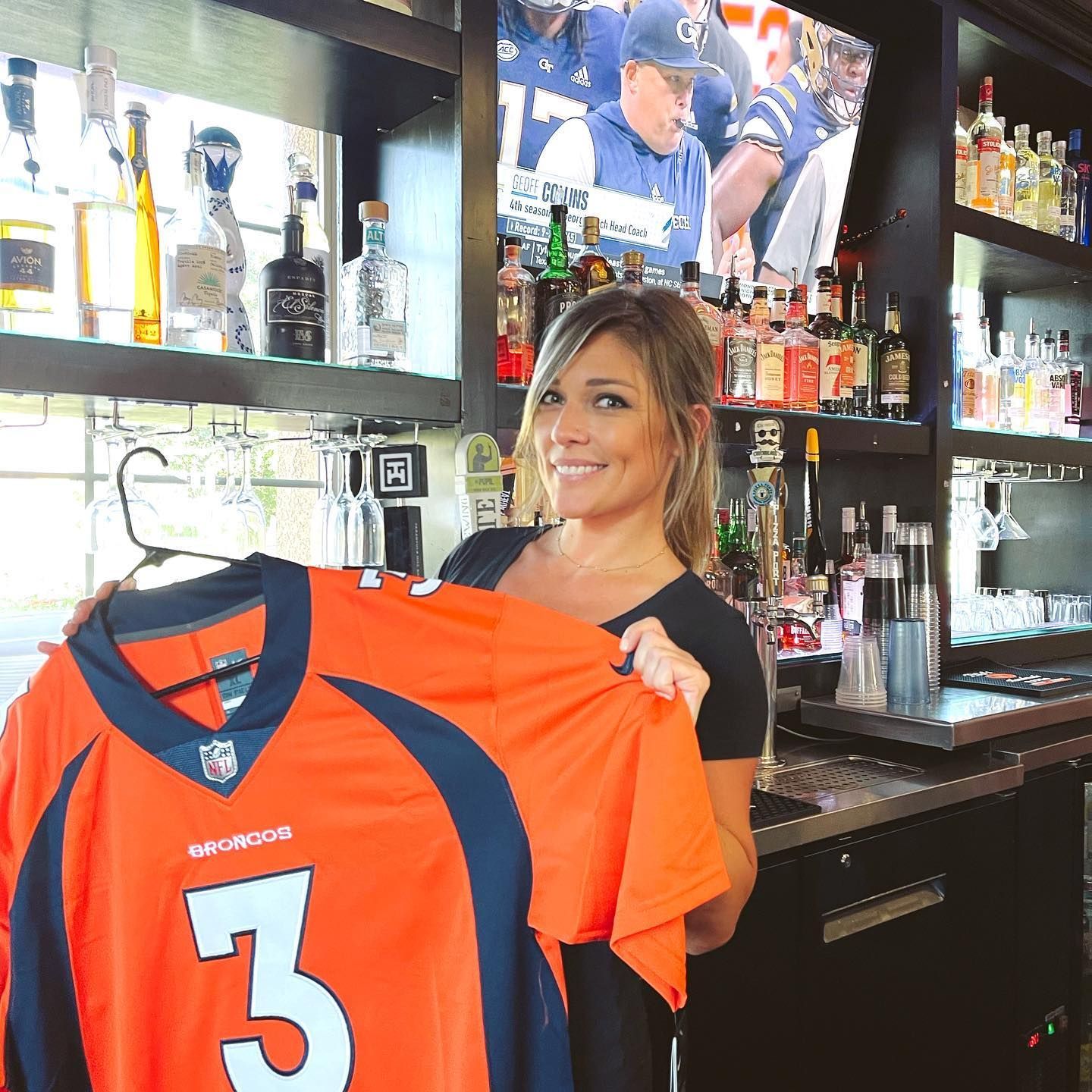 Woman holding a Denver Broncos jersey in a bar, TV in background with football game.