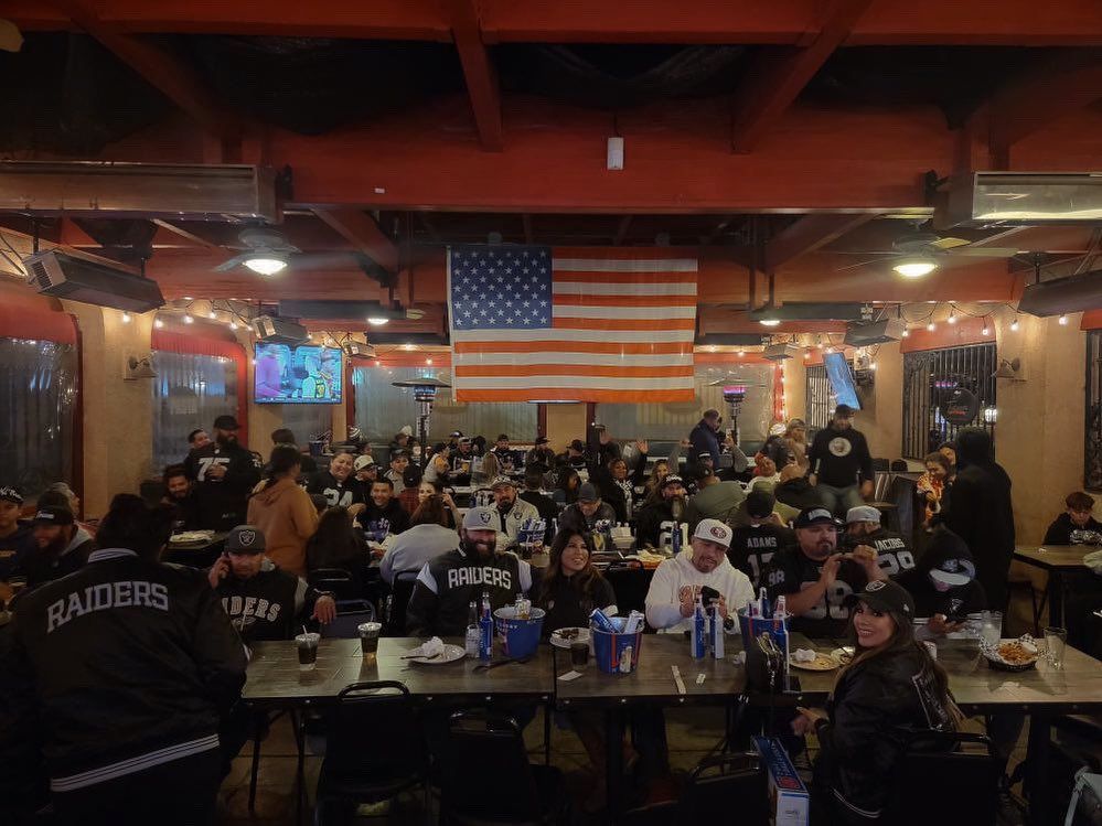 Bar packed with people, many in Raiders gear, watching TV; American flag hangs overhead.