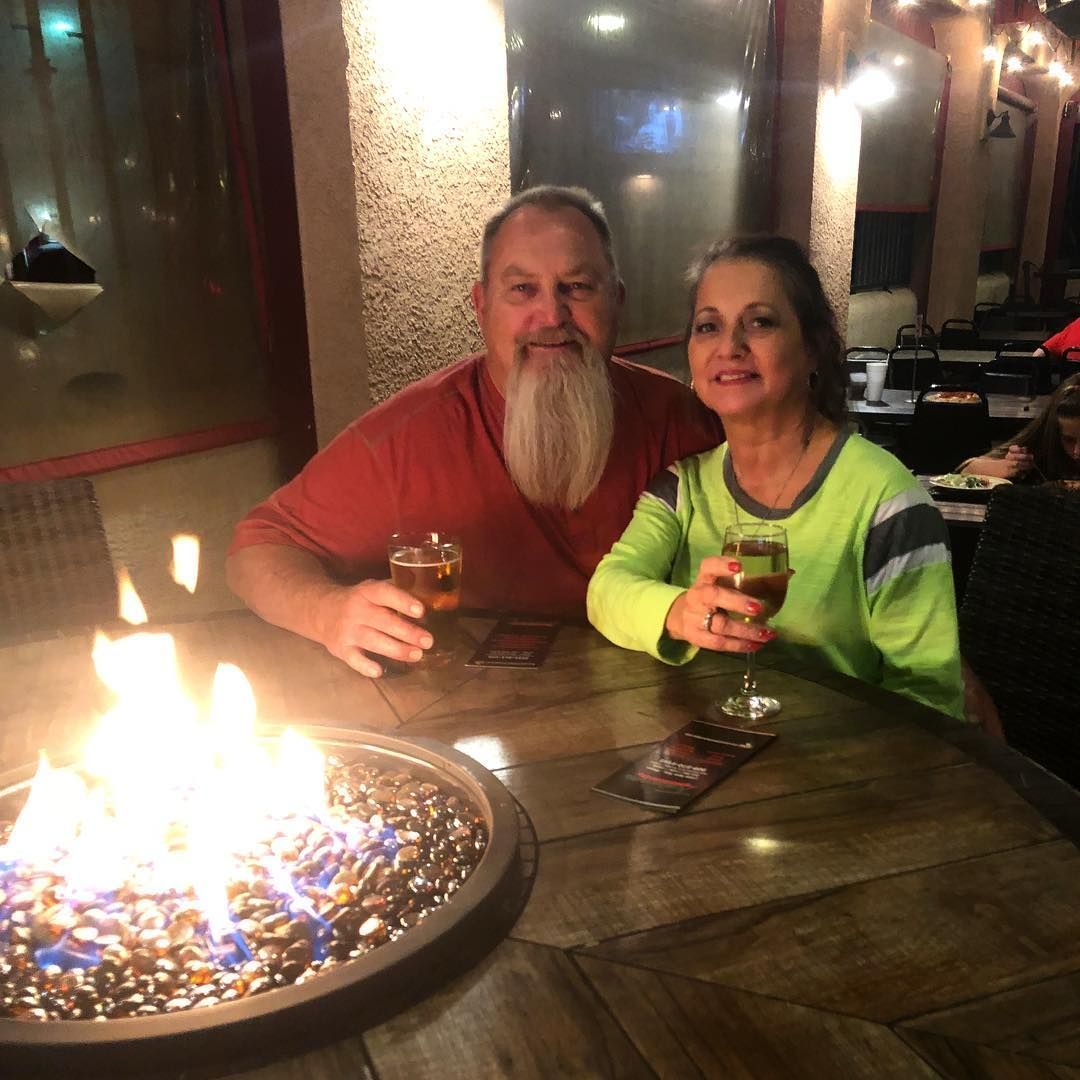 Couple toasting drinks by a fire pit. Man with a long beard, woman smiling. Outdoors, at a table.