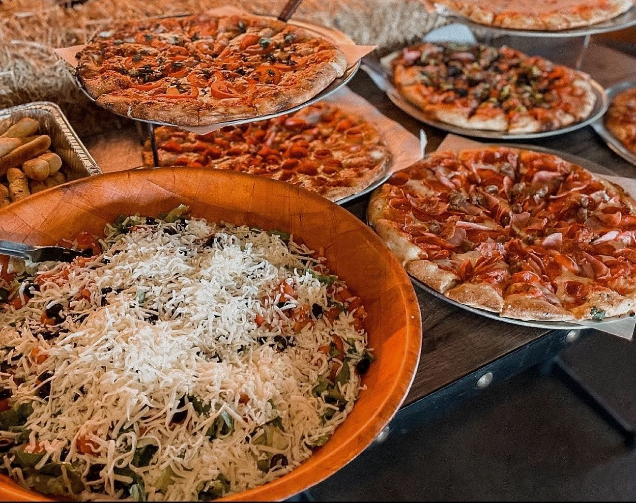 Table of food with multiple pizzas and a large bowl of salad; hay bales in background.
