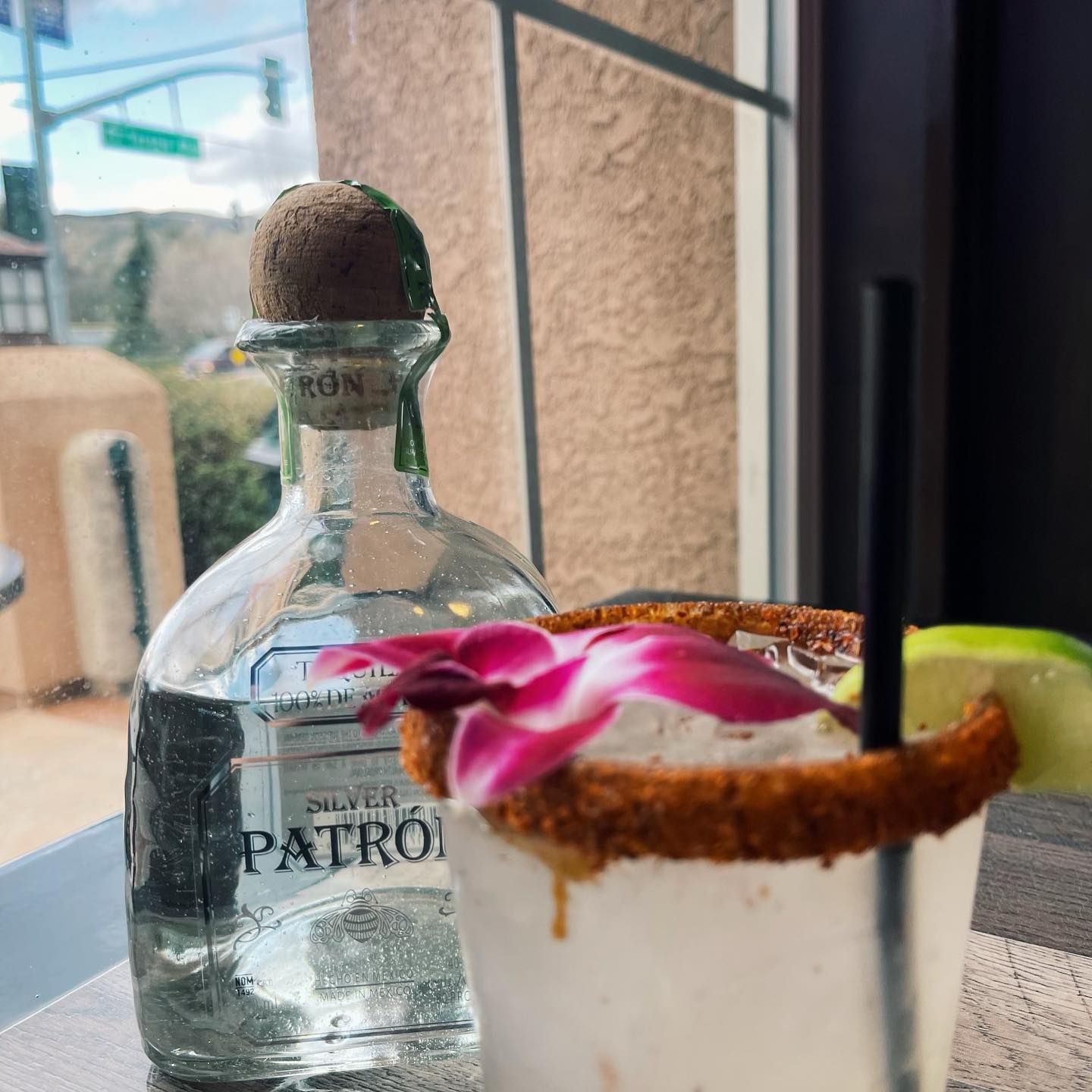 Bottle of Patron tequila next to a cocktail with a lime and flower garnish, sitting by a window.