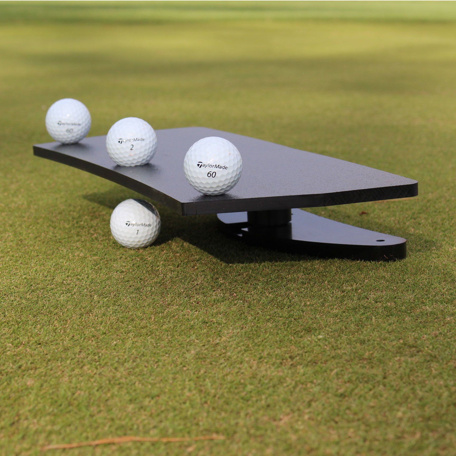 Black golf putting training aid with four golf balls on green grass.