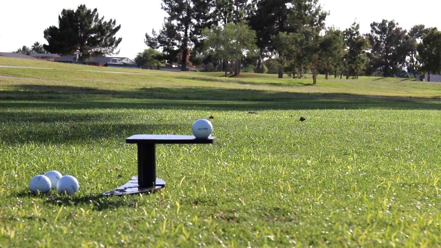 Golf ball on a tee, striking another ball, on green grass. Trees and sky in the background.