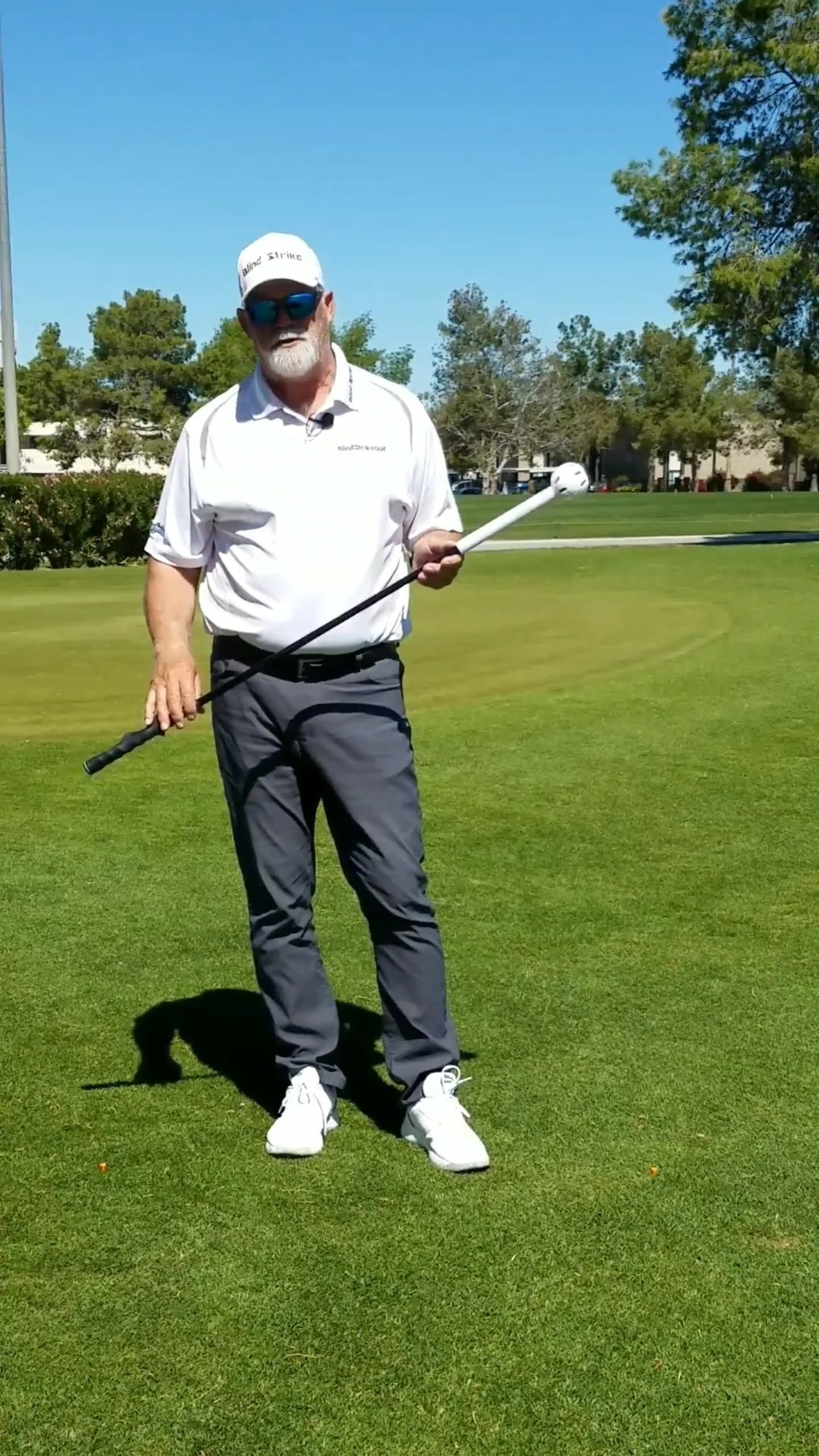 Man in white shirt and hat holding golf training aids on a green golf course under a blue sky.