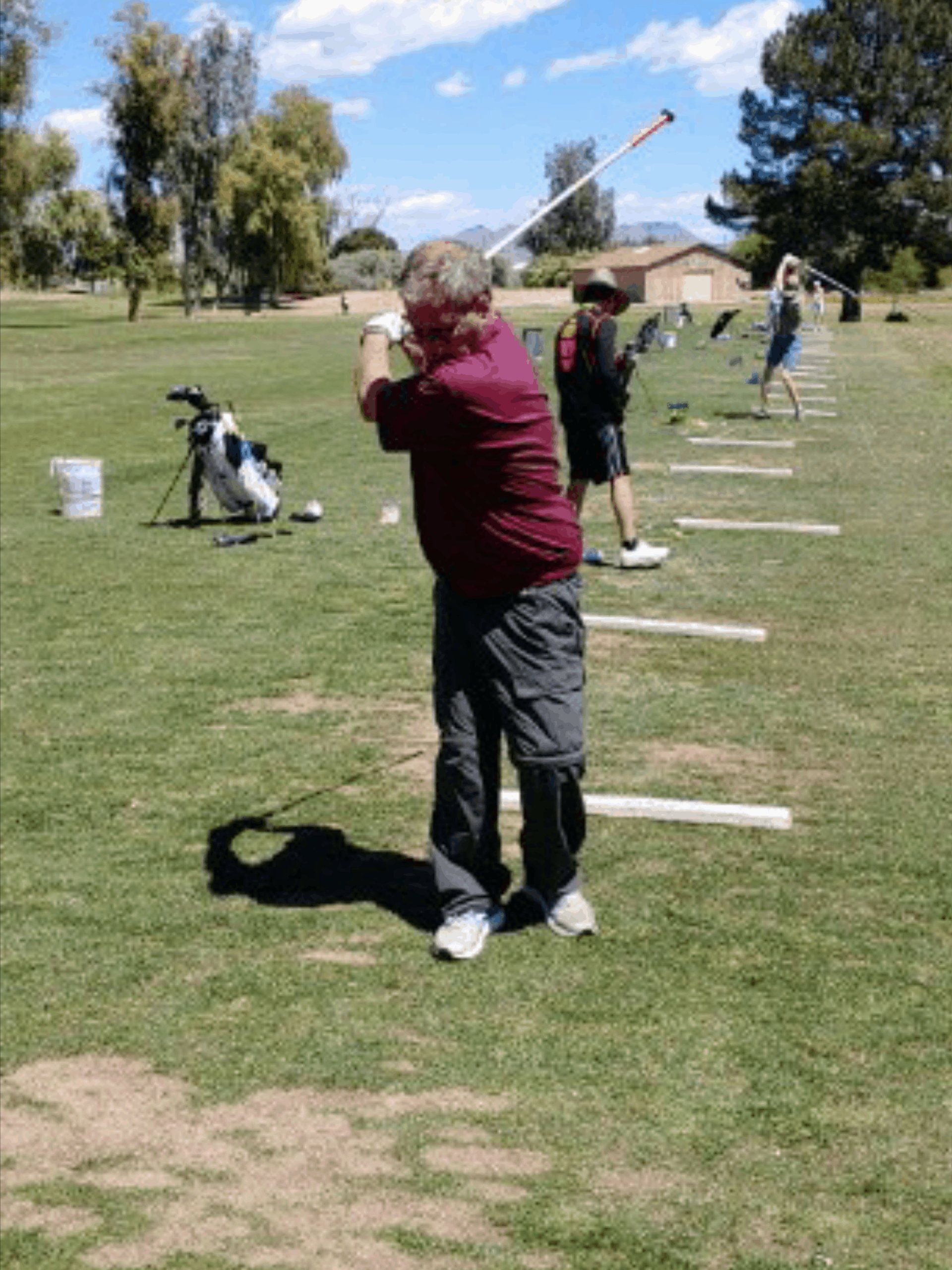 Man in maroon shirt swinging a golf club at a driving range under a sunny sky.
