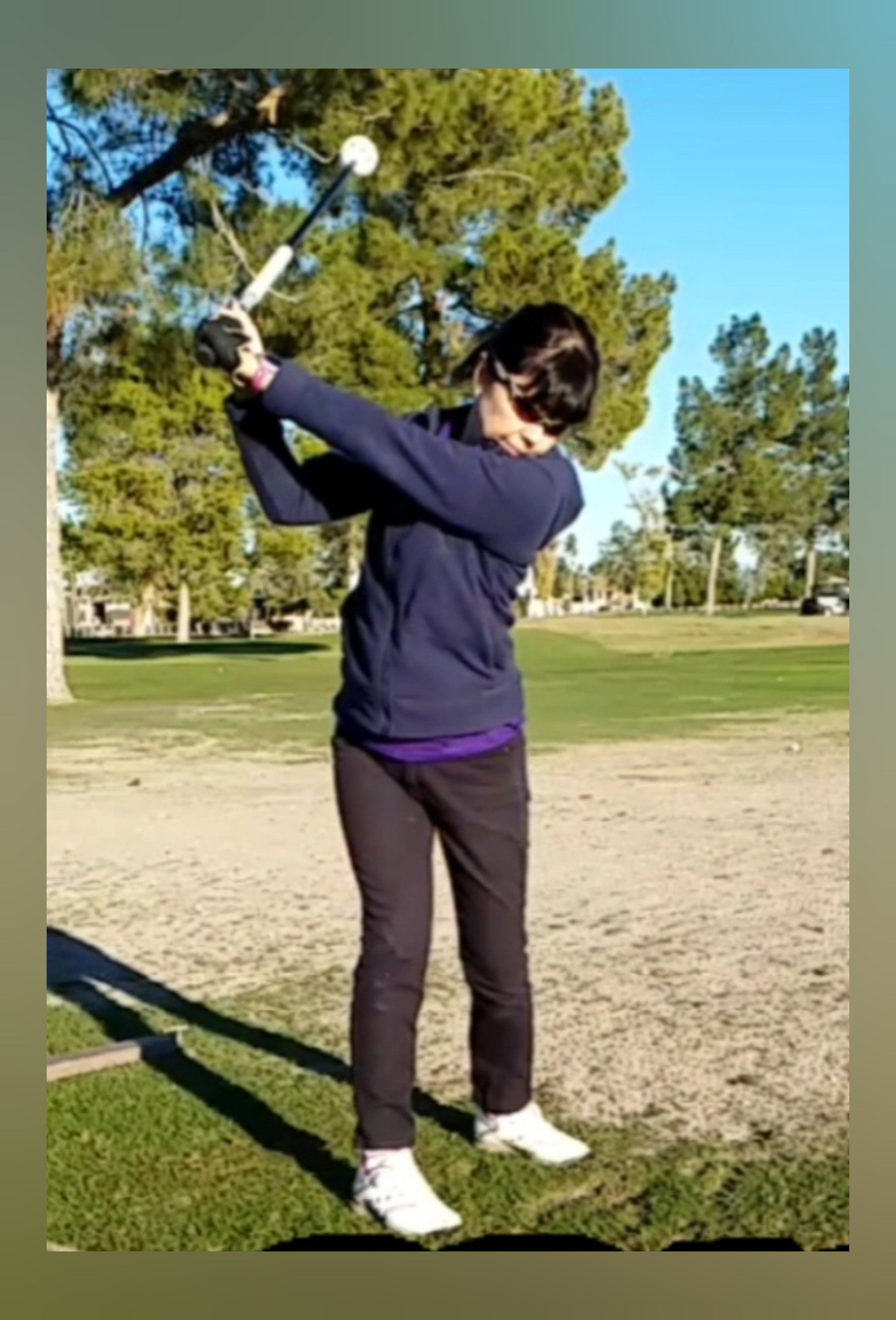 Woman in dark clothes swings a golf club at a course, trees and sky in the background.