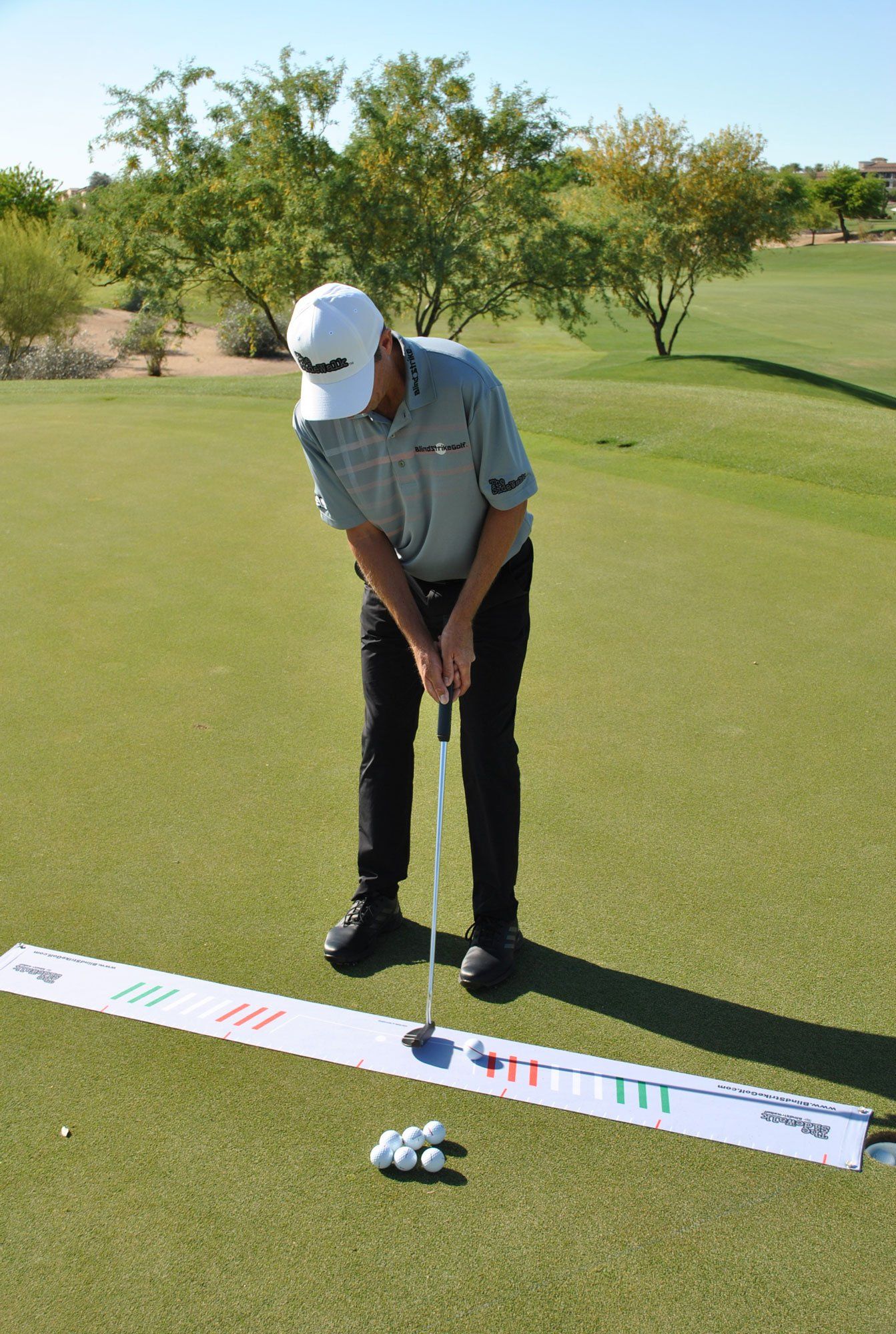 Golfer putting on green with training aid and golf balls. Sunny outdoor setting.