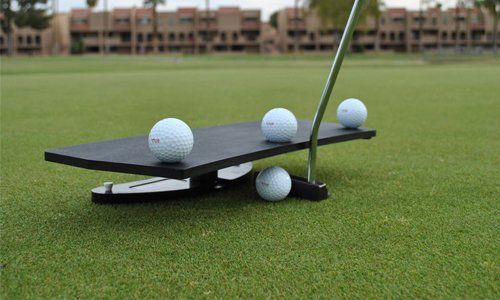 Golf putting training aid with four golf balls on a green, balanced on a black platform.