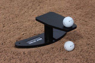 Black golf tee with golf ball on top, another ball beside it, on sand.