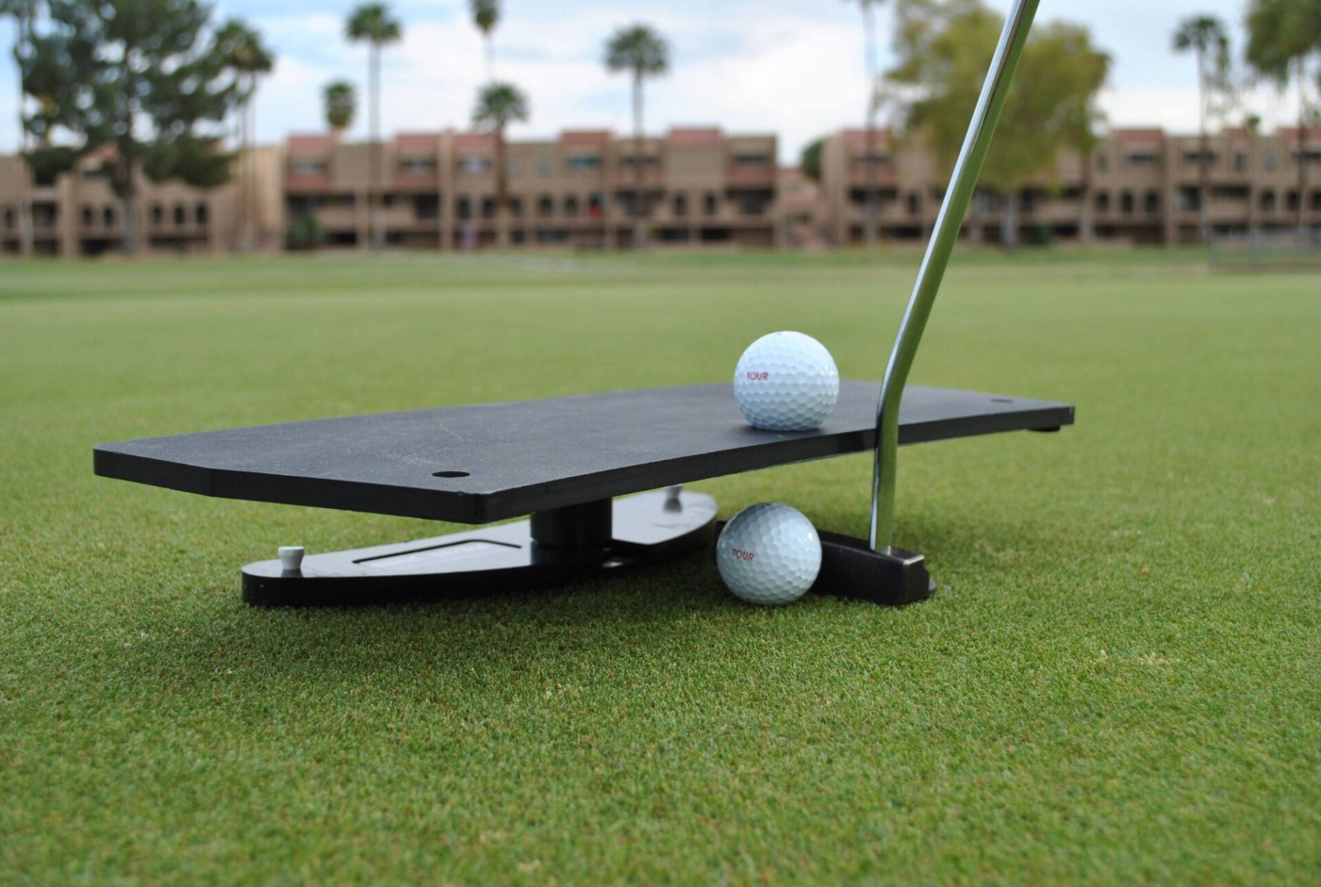 A putting training aid on a green with two golf balls; the aid holds one ball in place.