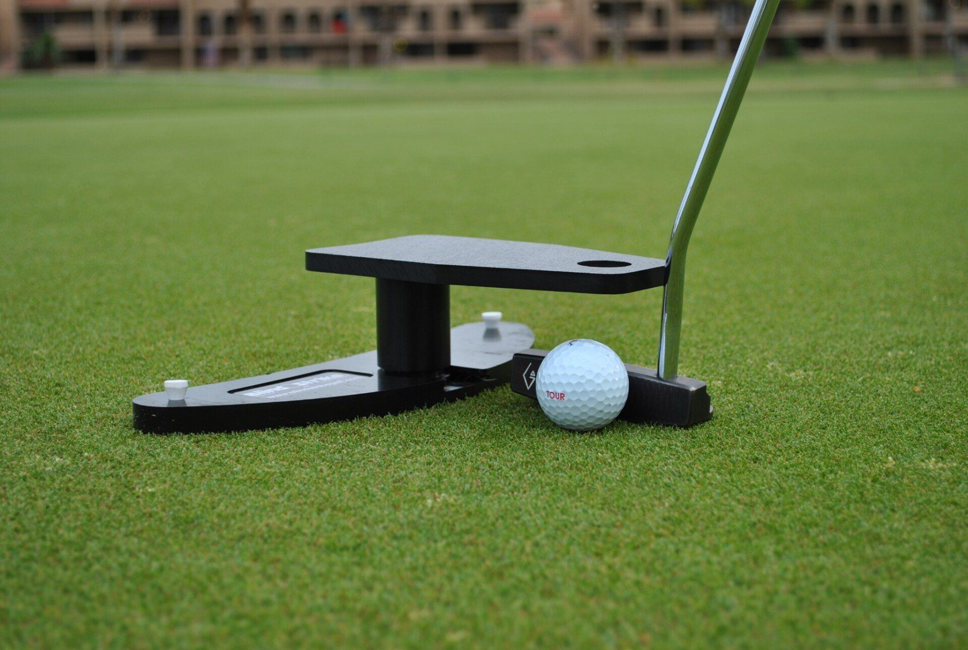 Golf ball near a putting aid on a green. The aid is black, with a golf club poised above the ball.