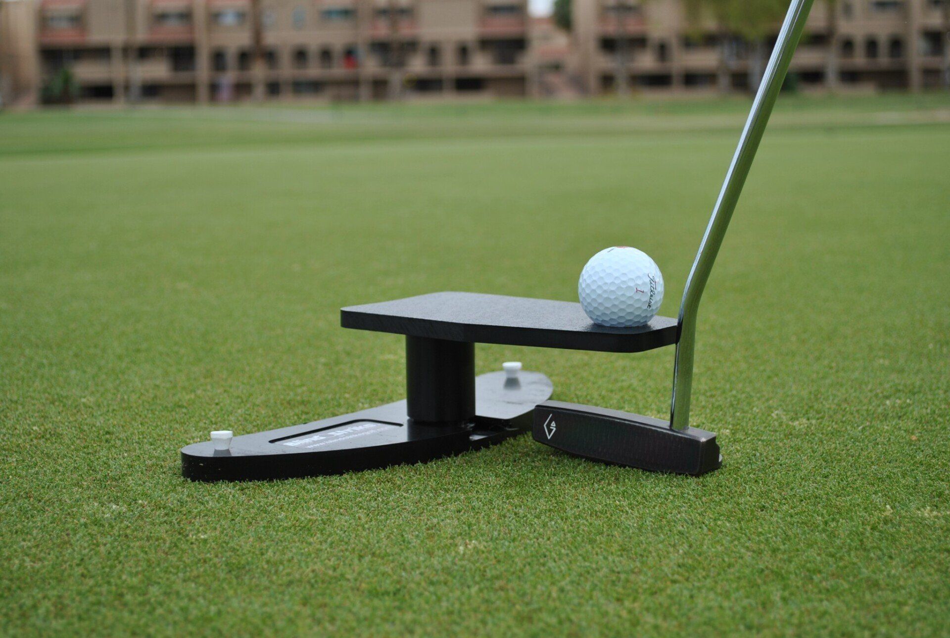 A golf ball sits on a black putting aid on a green. A putter rests beside the ball.