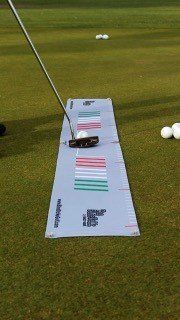 Golfer putts a ball on a putting mat with color-coded lines on a green. Balls and white markers are in background.