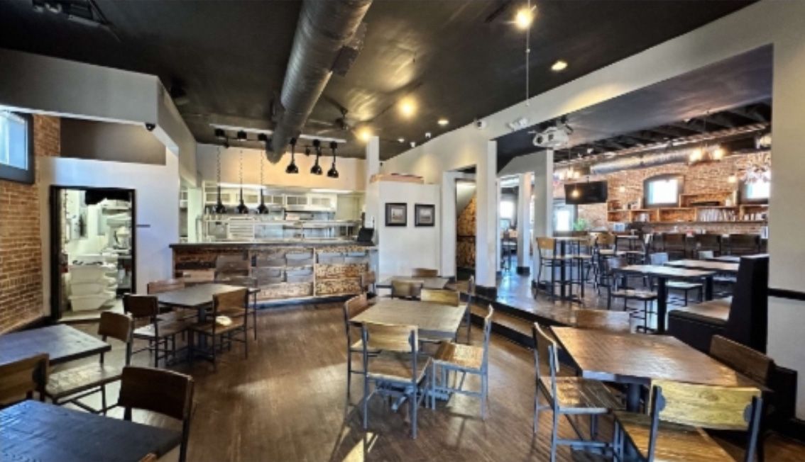 Restaurant interior with tables, chairs, bar, and kitchen area, featuring exposed brick, wood, and a dark ceiling.